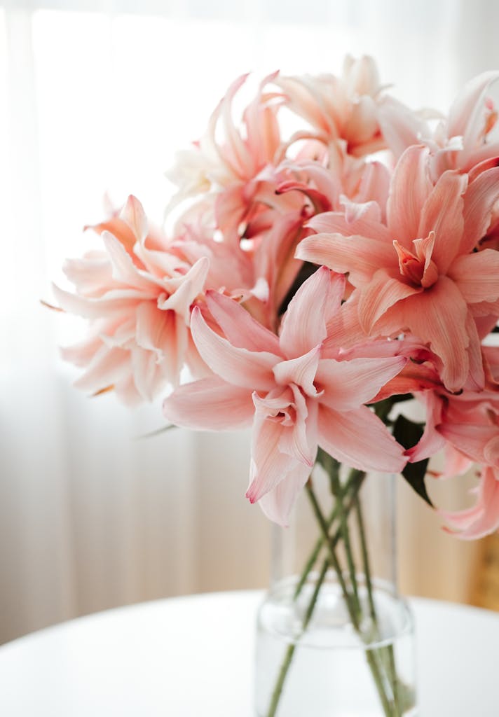 Delicate pink lilies arranged in a clear glass vase on a white table.